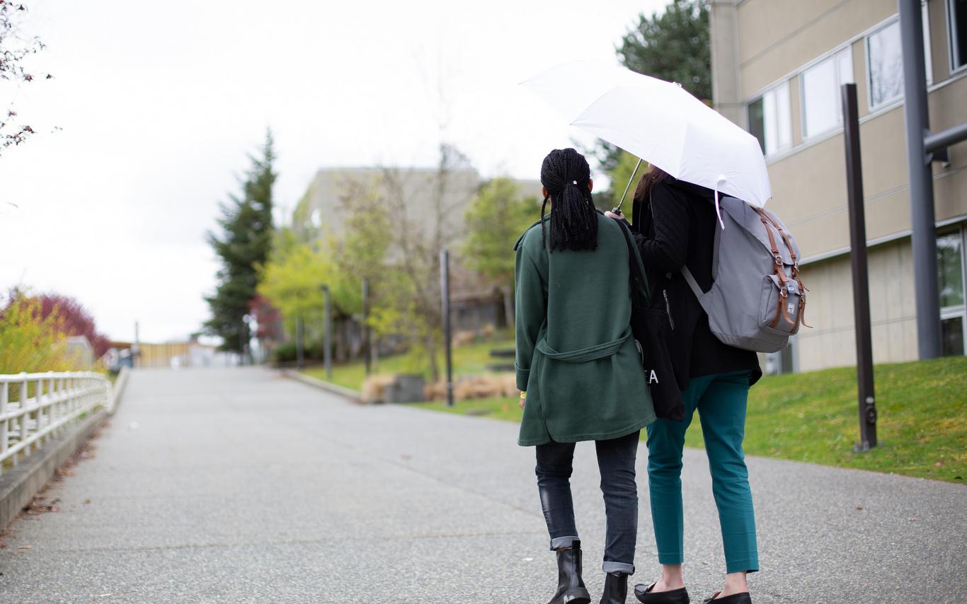 Two figures under one umbrella outside on campus.